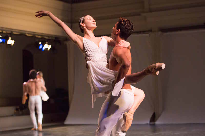 Two couples one in the foreground and another in the background. The men carry the women. The woman closest to the camera grins at her partner as she extends her right arm above her shoulder and extends her right foot parallel to the floor.